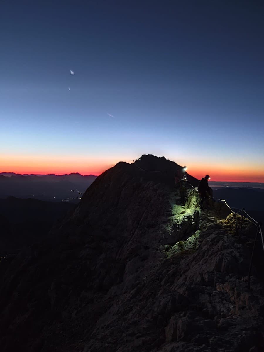 Hikers with headlamps ascending rocky mountain ridge at pre-dawn twilight.