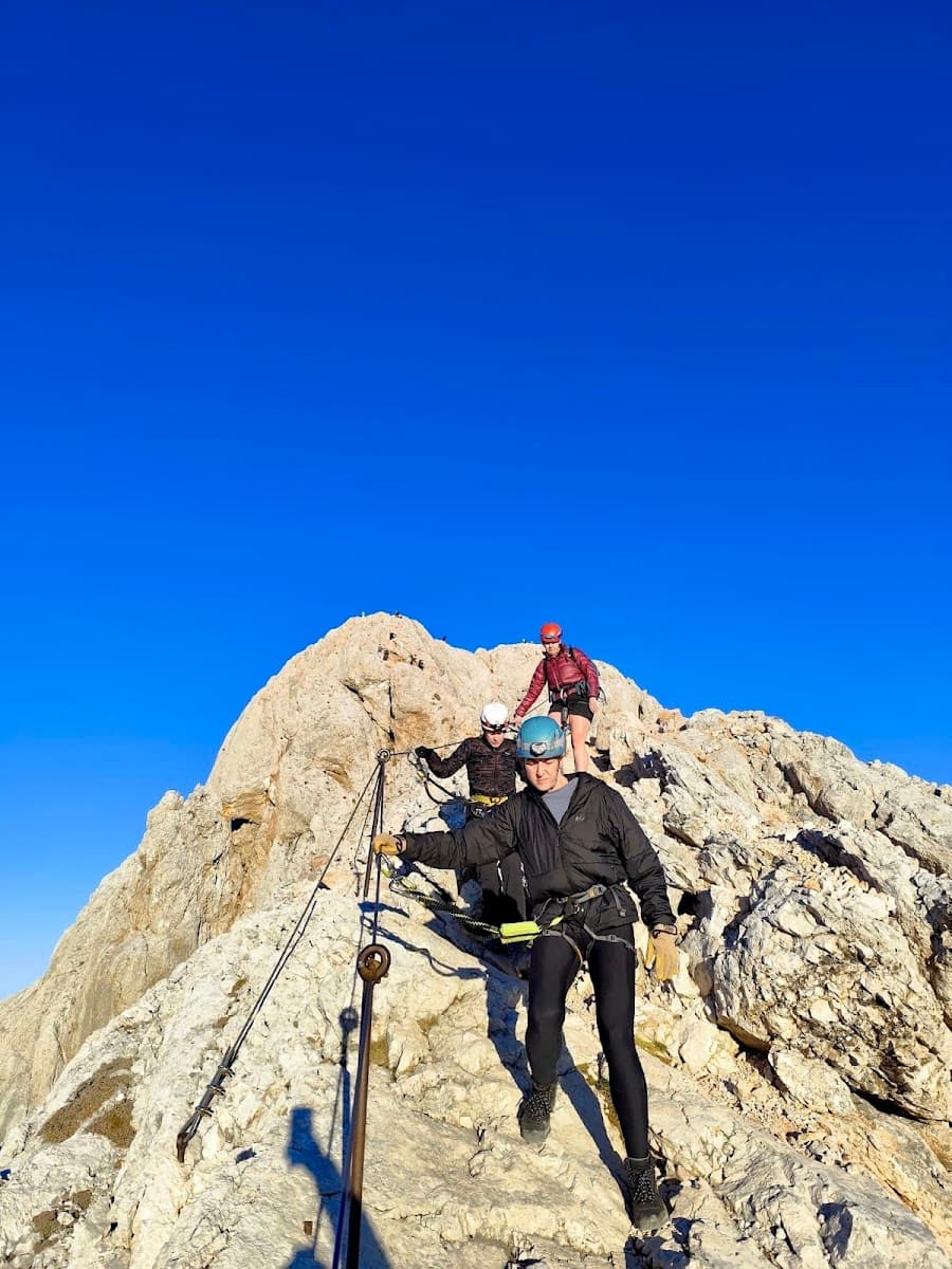 Hikers with helmets ascending steep, rocky mountain using fixed cables under a clear blue sky.