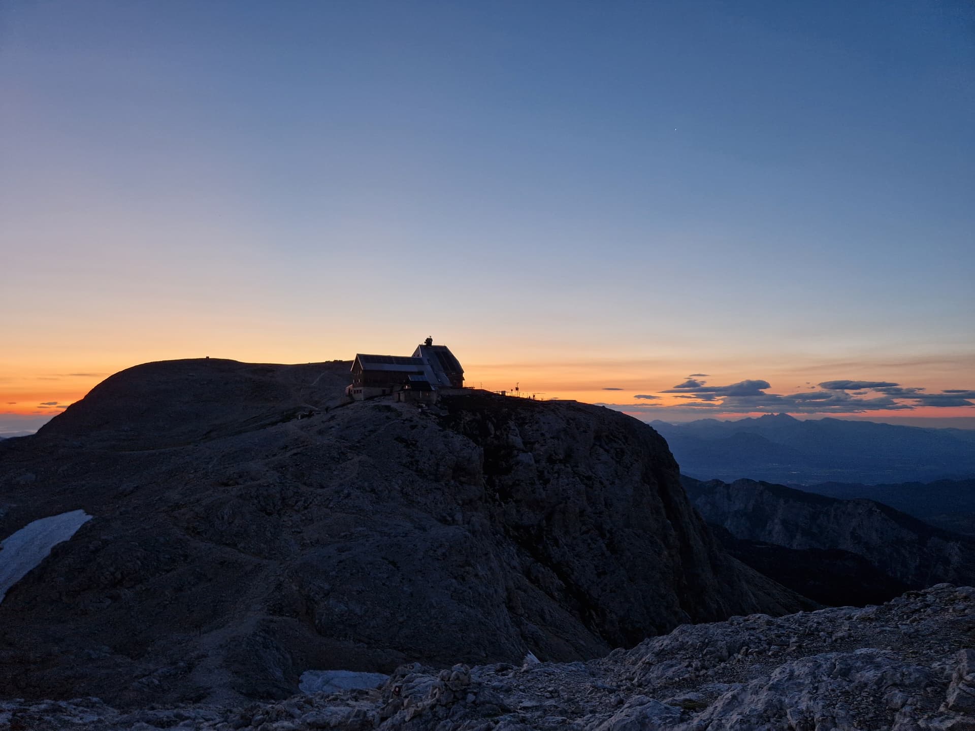 Mountain hut silhouetted on rocky peak at sunrise over distant mountain range