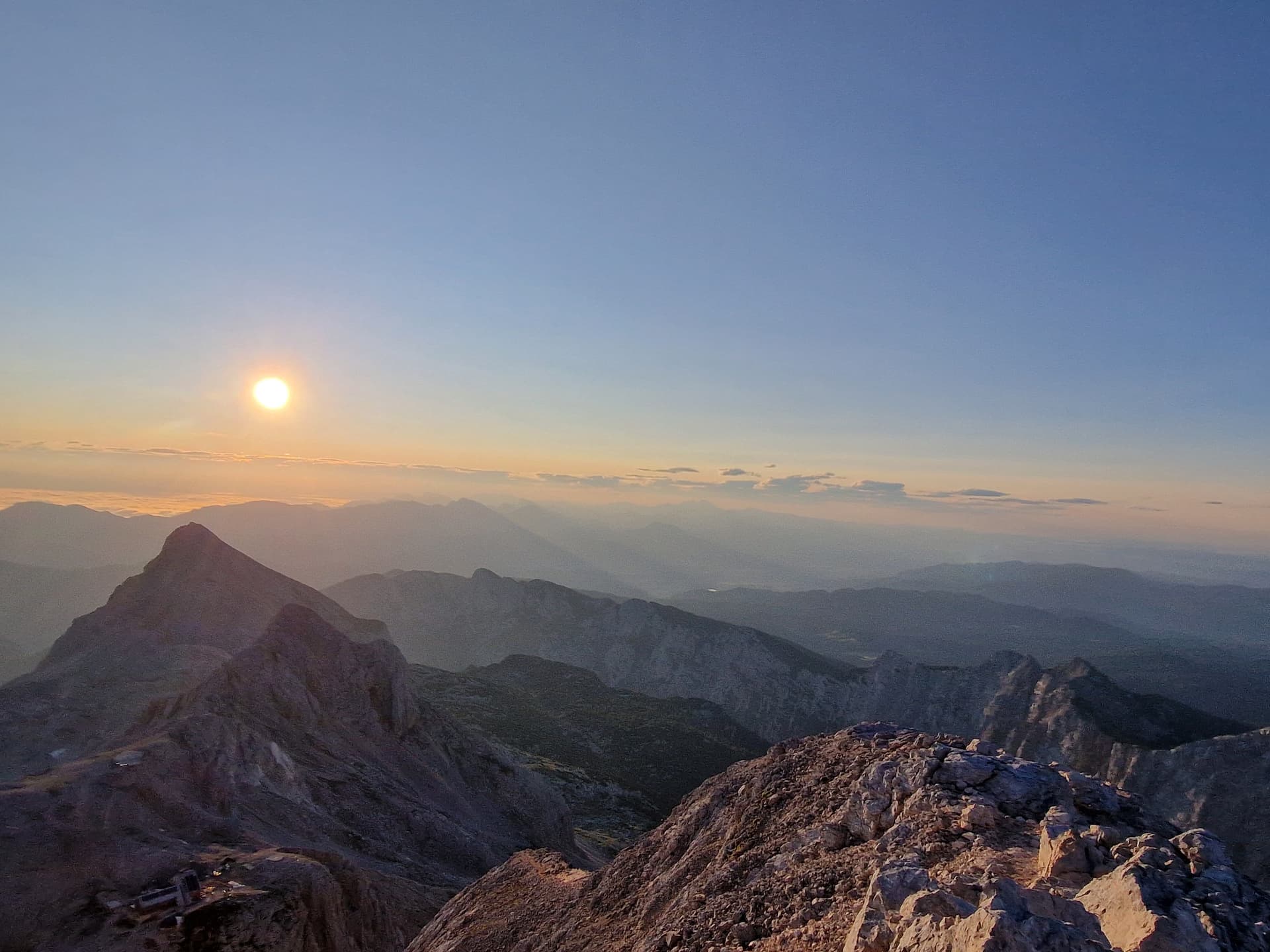 Rocky mountain summit overlooking layered peaks at sunrise with the sun low on the horizon.