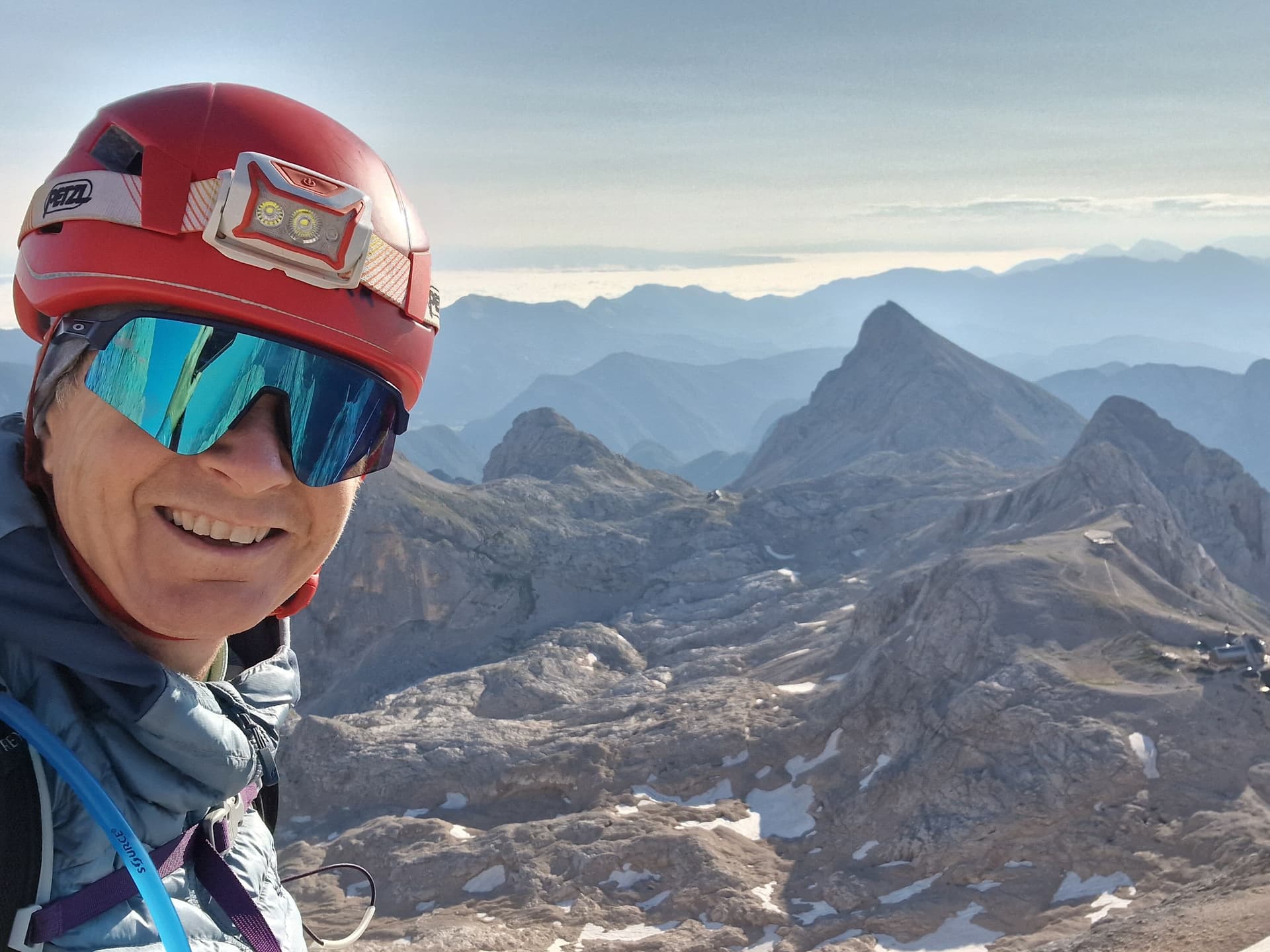 Hiker with helmet and sunglasses smiling above rocky mountains with patches of snow.