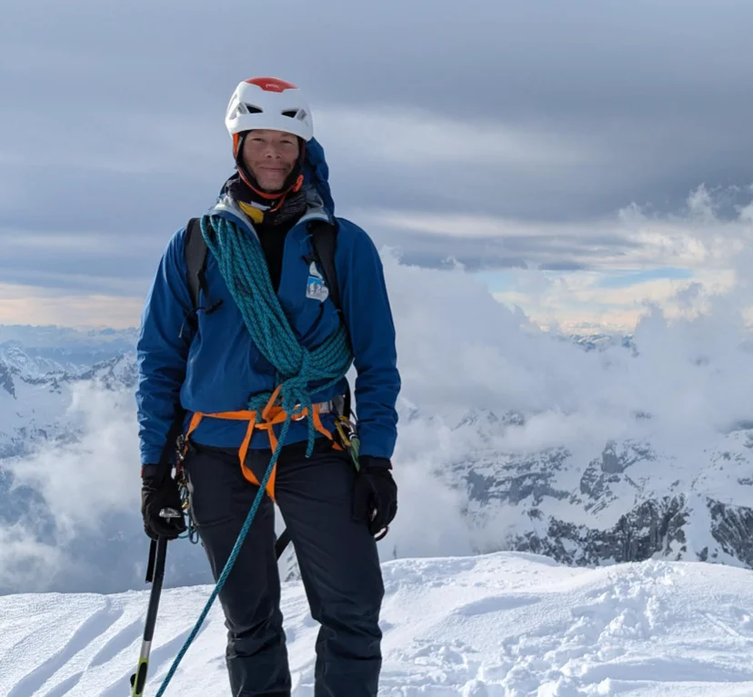 Mountaineer in climbing gear standing on a snow-covered mountain peak with clouds in the background.