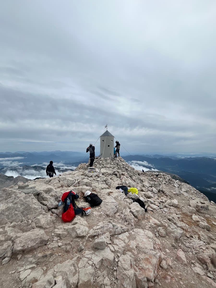 Hikers at Aljaž Tower on rocky mountain summit under cloudy sky