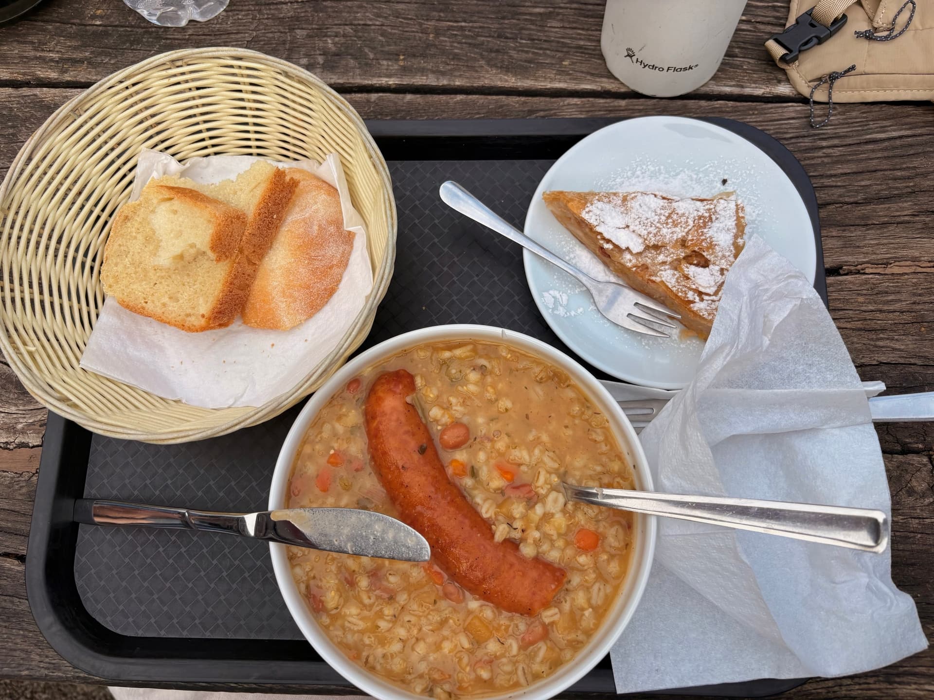 Hearty stew with a sausage, sliced bread in a basket, and pastry on a wooden table at a mountain hut