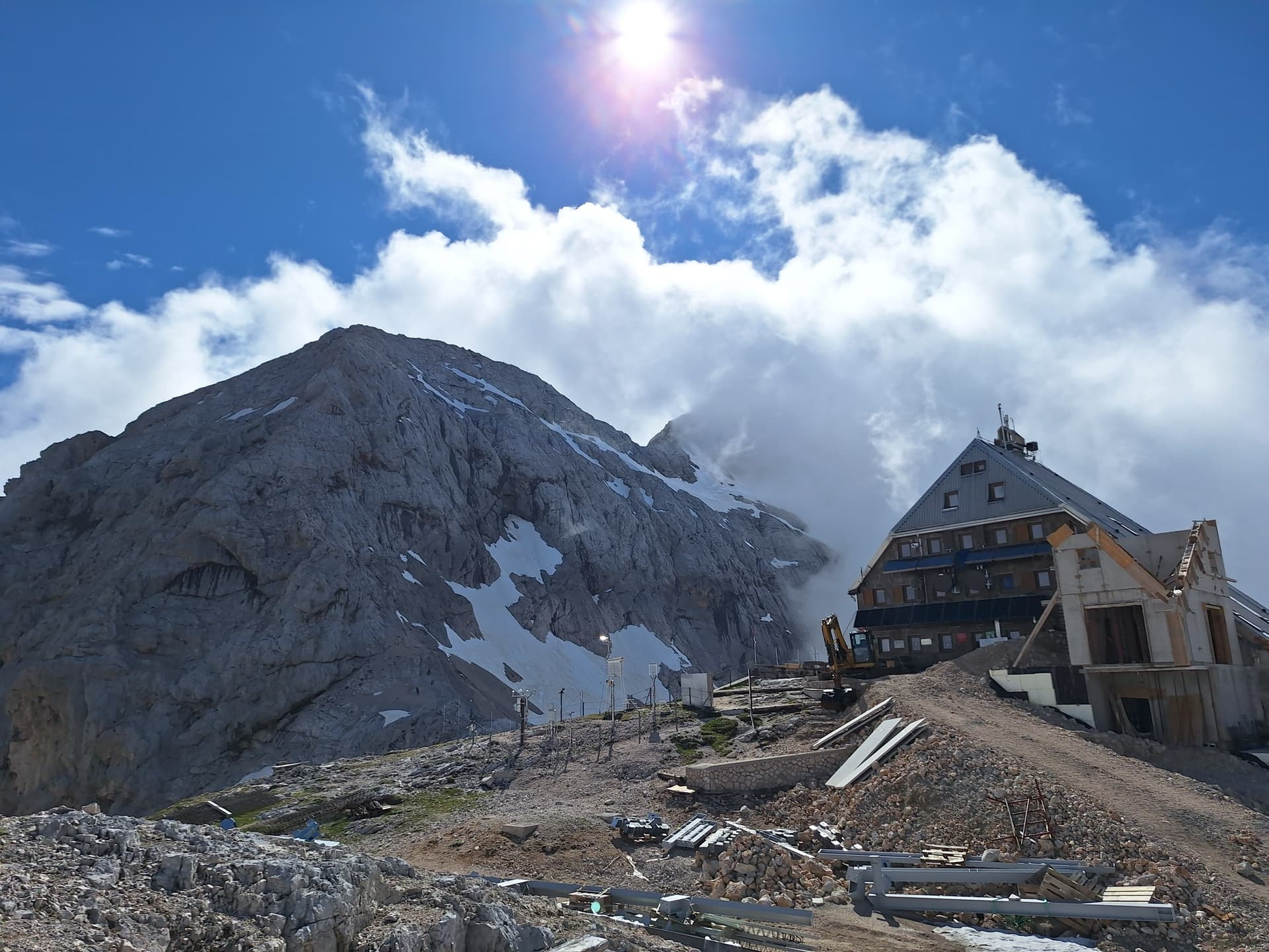 Mountain hut below snowy summit under bright sun and blue sky with clouds