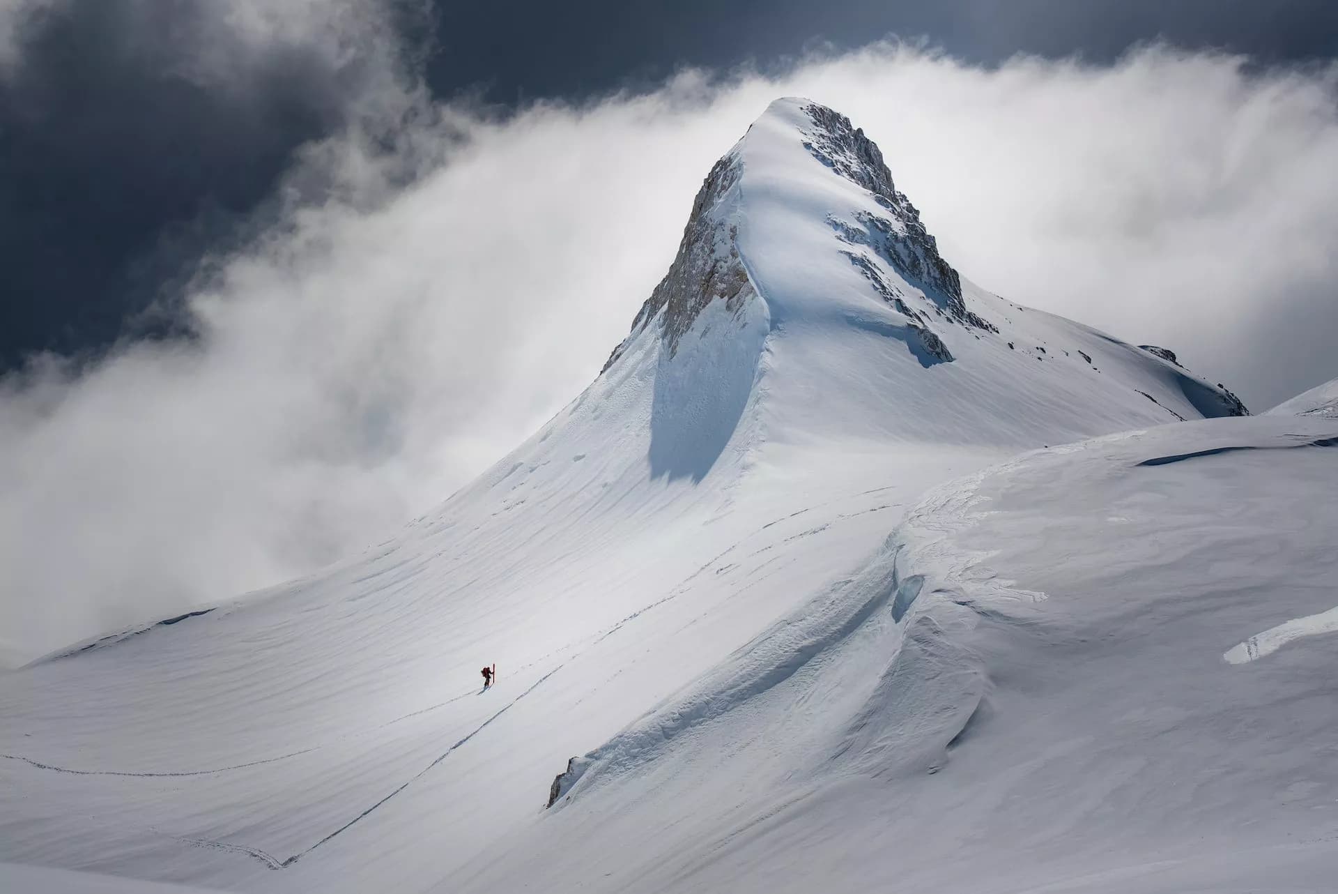 Ski touring below Kredarica peak on a vast, snow-covered mountain slope under cloudy sky.
