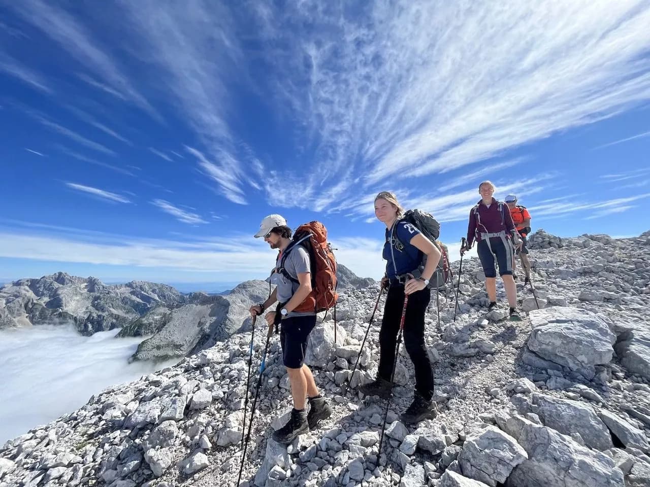 Hikers with backpacks traverse rocky terrain above the clouds with Julian Alps peaks in the background.